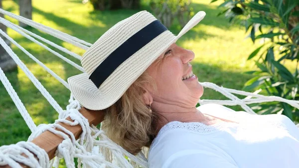 Seniorka v slamáku se šťastně usmívá, odpočívá na houpací síti a užívá si čerstvého vzduchu na terase kolem palem u moře. Koncept životního stylu seniorů — Stock fotografie