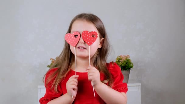 Ładna dziewczynka w czerwonej sukience z dwoma sercami na patyku zamyka nimi oczy. Koncepcja Walentynek. — Wideo stockowe