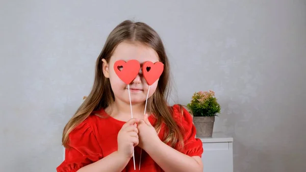 Jolie petite fille dans une robe rouge avec deux cœurs sur un bâton ferme les yeux avec eux. Concept de Saint-Valentin. — Photo