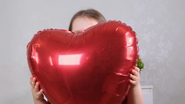 Маленькая милая девочка в красном платье с красными шарами в форме сердца в руках концепция Дня Святого Валентина — стоковое видео