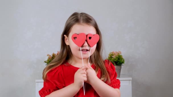 Hübsches kleines Mädchen in rotem Kleid mit zwei Herzen auf einem Stock schließt mit ihnen die Augen. Valentinstag-Konzept. — Stockvideo