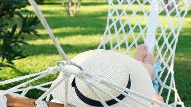 Hasır şapkalı bir kadın palmiye ağaçlarının etrafındaki bir hamakta dinleniyor ve huzur ve rahatlığın tadını çıkarıyor. Tatil kavramı