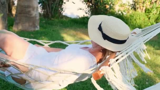 Ontspannen senior vrouw met strohoed glimlachend gelukkig ontspannen op een hangmat genieten van de frisse lucht op het terras rond de palmbomen in de buurt van de zee. Oudere burger levensstijl concept — Stockvideo