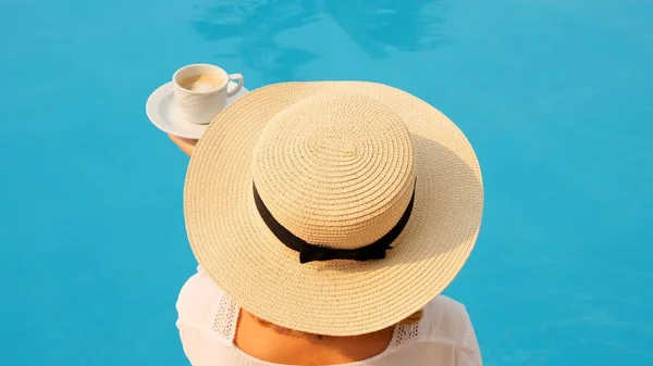 Жінка 50 років поснідала біля басейну в солом'яному капелюсі в білій сукні. жінка сидить біля басейну з чашкою кави. доброго ранку і денного планування — стокове фото