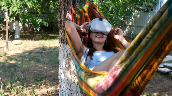 Happy smiling woman wearing virtual reality glasses rests in a hammock near a tree, imagining a dream come true, an invented world. The concept of getting experience using VR headset glasses of — Fotografia de Stock