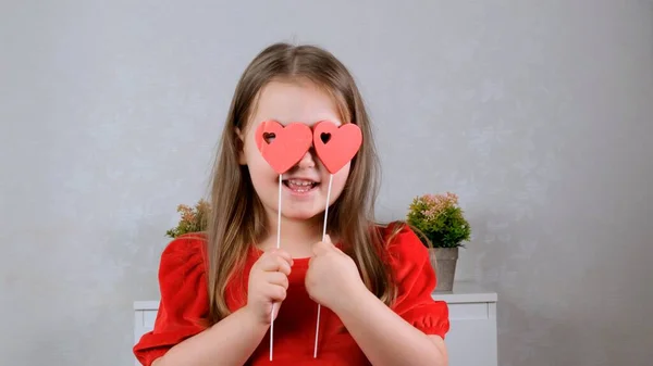 Gadis kecil cantik dengan gaun merah dengan dua hati pada tongkat menutup matanya dengan mereka. Valentines konsep hari. — Stok Foto