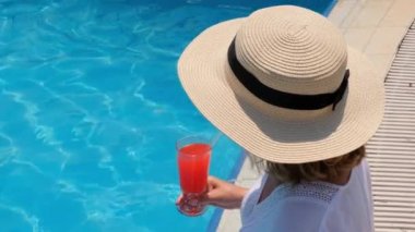 Hasır şapkalı sarışın kadın güneşli bir yaz gününde yüzme havuzunda turuncu kokteyl içiyor. Tatil konsepti, hepsi dahil.