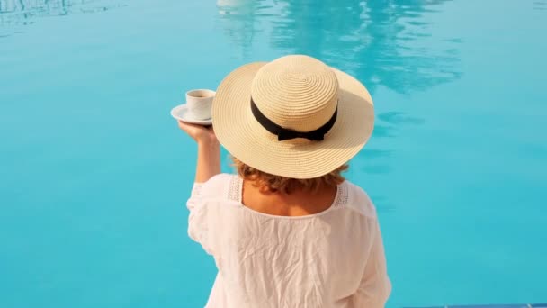 Fêmea com um chapéu de palha vestindo um vestido branco relaxante junto à piscina com uma xícara de café, conceito de férias, bom dia. mulher sentada à beira da piscina — Vídeo de Stock
