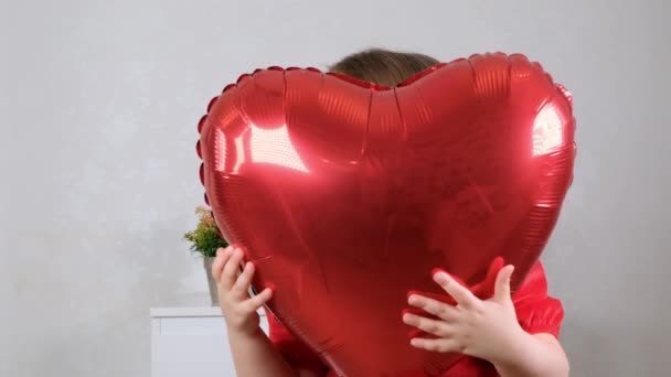 Little cute girl in a red dress hides behind a red heart shaped balloon. valentines day concept — Stock Video