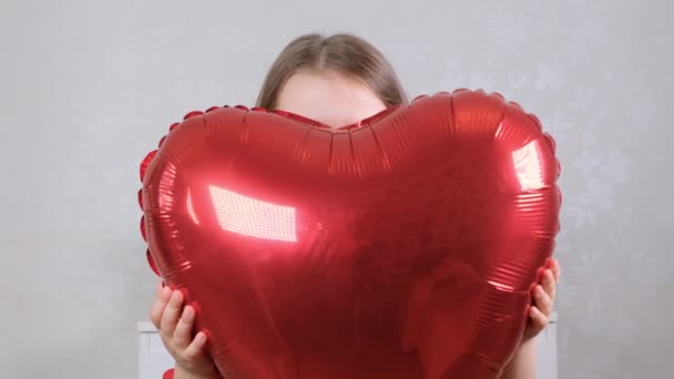 Маленькая милая девочка в красном платье прячется за красным шаром в форме сердца. День святого Валентина — стоковое видео