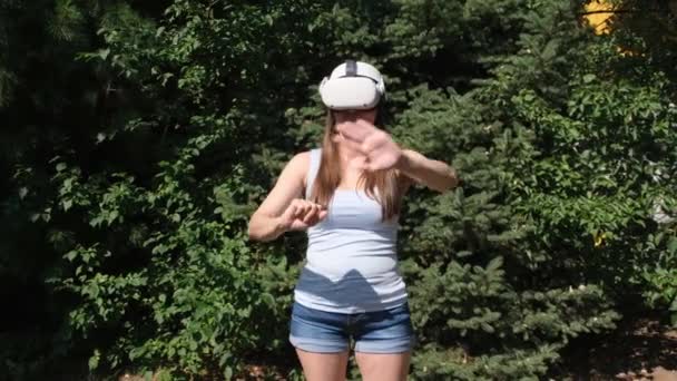Pensive Female in virtual reality glasses controls the device with his hand against the background of green trees. getting experience using VR headset glasses of virtual reality — Vídeo de Stock