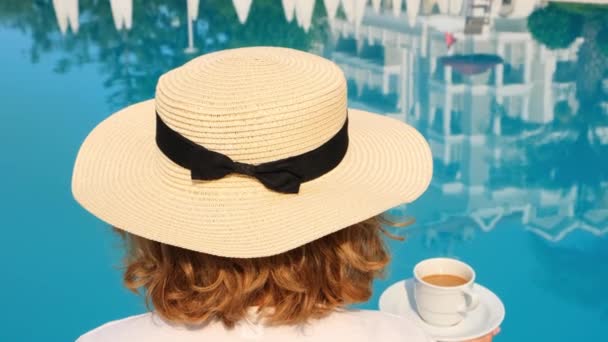 Θηλυκό σε ένα ψάθινο καπέλο κρατά ένα φλιτζάνι καφέ στο χέρι της πάνω από μια μπλε πισίνα έννοια του καλημέρα και την ημέρα του σχεδιασμού, χαλάρωση και απόλαυση, πίσω όψη — Αρχείο Βίντεο