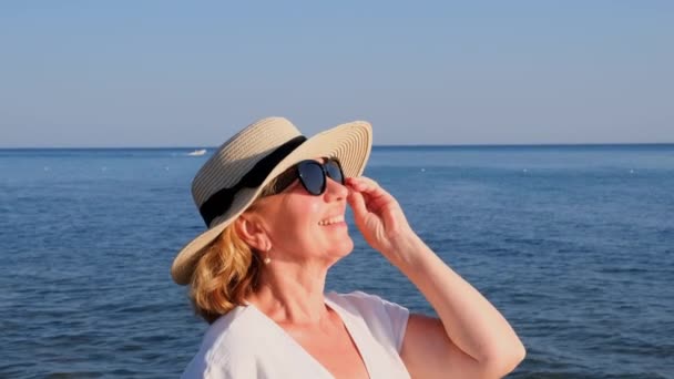 Portret zadowolonej 50-letniej kobiety w słomkowym kapeluszu i okularach przeciwsłonecznych cieszącej się słońcem nad błękitnym morzem. Lato, wakacje, wakacje, aktywni emeryci — Wideo stockowe