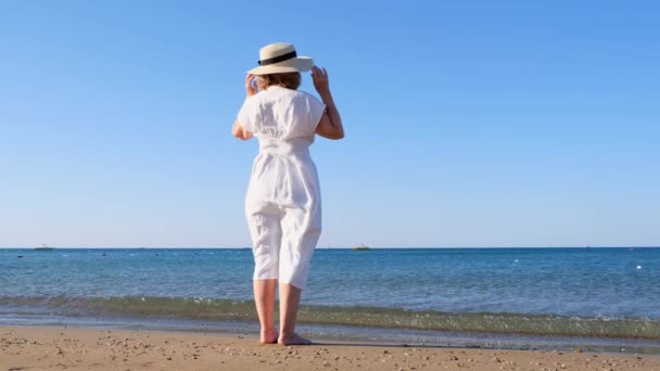 Kobieta w słomkowym kapeluszu i białej sukience spaceruje wzdłuż wybrzeża i podziwia błękitne morze w słoneczny letni dzień, ciesząc się wolnością i relaksem. Koncepcja życia codziennego seniora. — Wideo stockowe