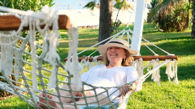 50-60 yaşlarında hasır şapkalı bir kadın palmiye ağaçlarının etrafındaki bir hamakta dinleniyor ve huzur ve rahatlığın tadını çıkarıyor. Emeklilik ve yaşam tarzı kavramı