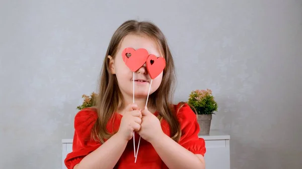 Jolie petite fille dans une robe rouge avec deux cœurs sur un bâton ferme les yeux avec eux. Concept de Saint-Valentin. — Photo