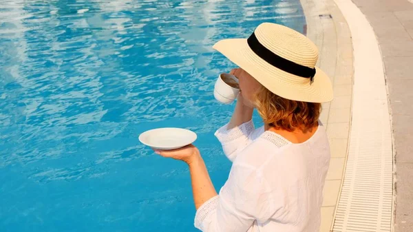 Крупним планом жінка у віці 50-55 років в солом'яному капелюсі п'є з чашки кави поруч з блакитним басейном, розкішний хороший ранок, початок дня — стокове фото
