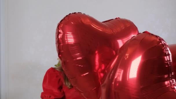 Srandovní holčička v červených šatech skrývající se za třemi balónky ve tvaru srdce na Valentýna. rodinná láska. dárky na svátky. štěstí a polibky — Stock video