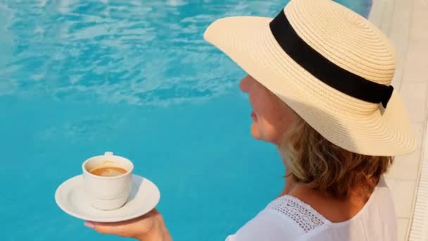 Femmina 50 anni con un cappello di paglia indossa un abito bianco rilassante a bordo piscina con una tazza di caffè, concetto di vacanza, buongiorno. donna seduta vicino alla piscina. — Video Stock