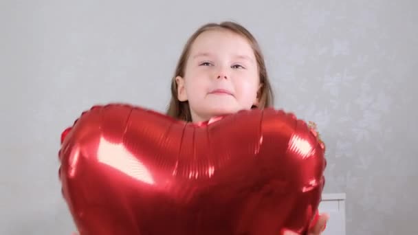 Маленькая милая девочка в красном платье с красными шарами в форме сердца в руках концепция Дня Святого Валентина — стоковое видео