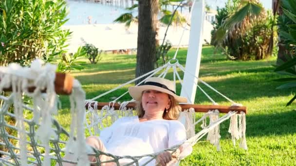 Funny senior woman in a straw hat smiling happy relaxing on a hammock enjoying the fresh air on the terrace around the palm trees near the sea. Senior citizen lifestyle concept — Αρχείο Βίντεο