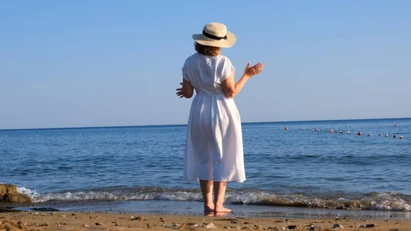 Зрелая женщина в соломенной шляпе и белом платье наслаждается солнцем, гуляя вдоль синего побережья моря в солнечный летний день, наслаждаясь свободой и отдыхом. Концепция повседневной жизни пожилых людей — стоковое фото
