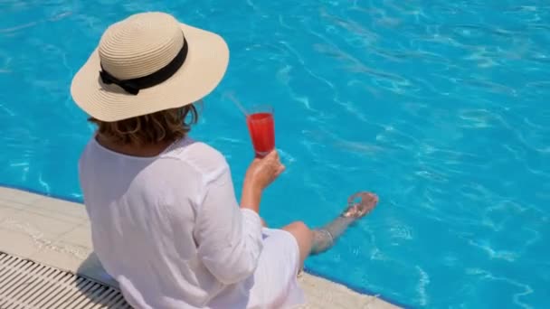 Mulher em um chapéu de palha relaxa perto da piscina, bebe um coquetel rosa e balança as pernas na água. Conceito de humor verão — Vídeo de Stock