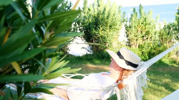 Senior woman in straw hat smiling happy relaxing on a hammock enjoying the fresh air on the terrace around palm trees near the sea. Senior citizen lifestyle concept — Stockvideo