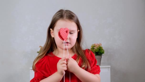 Grappig klein meisje in een rode jurk met twee harten op een stok sluit haar ogen met hen. Valentijnsdag concept. — Stockvideo