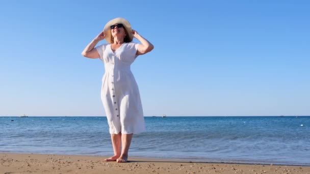 Mujer mayor en gafas de sol con sombrero de paja y vestido blanco camina a lo largo de la costa azul del mar en un día soleado de verano, disfrutando de la libertad y la relajación. El concepto de una vida típica de los pensionistas — Vídeos de Stock