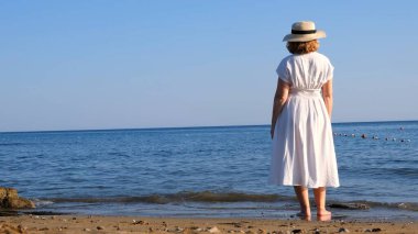 Hasır şapkalı ve beyaz elbiseli bir kadın deniz kıyısında yürüyor ve güneşli bir yaz gününde mavi denize hayran, özgürlüğün ve rahatlığın tadını çıkarıyor. Yaşlılar için günlük yaşam kavramı.