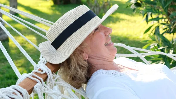 Ženy ve věku 50-60 let v slamáku odpočívají v houpací síti kolem palem a užívají si klidu a odpočinku. Koncept odchodu do důchodu a životního stylu — Stock fotografie