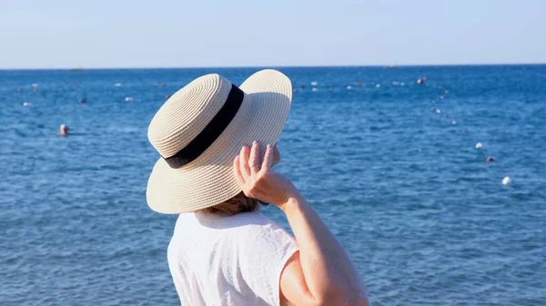 Πίσω όψη μιας γυναίκας με ψάθινο καπέλο και λευκό φόρεμα που κοιτάζει τη γαλάζια θάλασσα, από κοντά. Η έννοια της χαλάρωσης, ανάπαυσης, αντηλιακό — Φωτογραφία Αρχείου