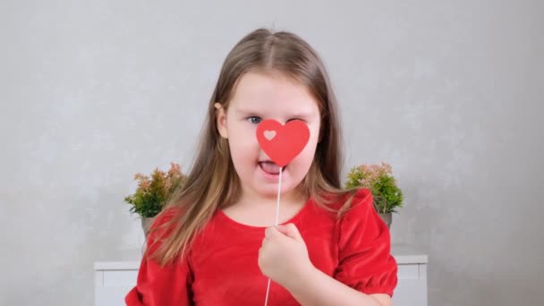 Funny Little girl in a red dress with a heart on a stick closes their eyes. Valentines day concept. — Stock Video