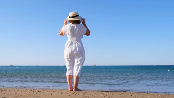 Güneşli bir yaz gününde mavi deniz kıyısında yürürken hasır şapkalı ve beyaz elbiseli olgun bir kadının arkasına bakmak özgürlük ve rahatlığın tadını çıkarıyor. Emeklilerin tipik hayatı kavramı. — Stok video