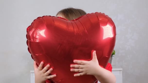 Маленька мила дівчинка в червоній сукні ховається за червоною кулею у формі серця. концепція дня валентинки — стокове відео
