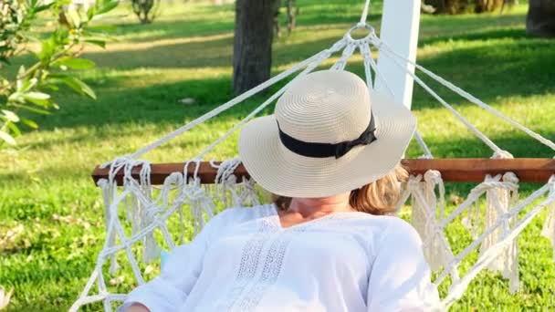 一位戴着草帽笑着笑着的有趣的老妇人坐在吊床上，享受着海边棕榈树周围的露台上的新鲜空气。老年公民生活方式概念 — 图库视频影像
