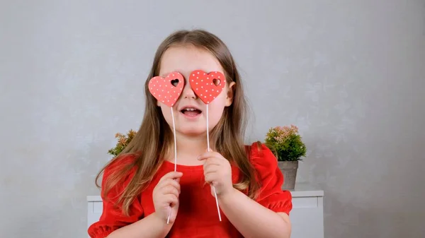 Jolie petite fille dans une robe rouge avec deux cœurs sur un bâton ferme les yeux avec eux. Concept de Saint-Valentin. — Photo