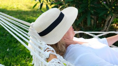 Hasır şapkalı bir kadın palmiye ağaçlarının etrafındaki bir hamakta dinleniyor ve huzur ve rahatlığın tadını çıkarıyor. Tatil kavramı