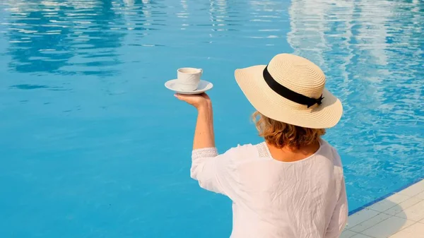 Жінка в солом'яному капелюсі в білій сукні, що розслабляється біля басейну з чашкою кави, концепцією відпустки, добрим ранком. жінка сидить біля басейну — стокове фото