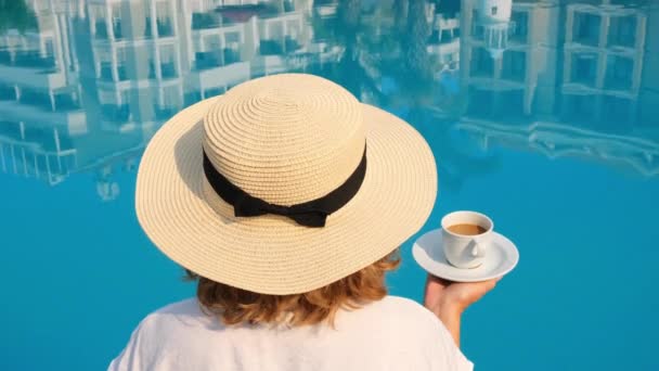 Mujer de 50 años con un sombrero de paja con un vestido blanco relajante junto a la piscina con una taza de café, concepto de vacaciones, buenos días. mujer sentada junto a la piscina. — Vídeos de Stock