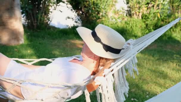 一位头戴草帽的老年妇女笑着轻松地坐在吊床上，享受着海边棕榈树周围的露台上新鲜的空气。老年公民生活方式概念 — 图库视频影像
