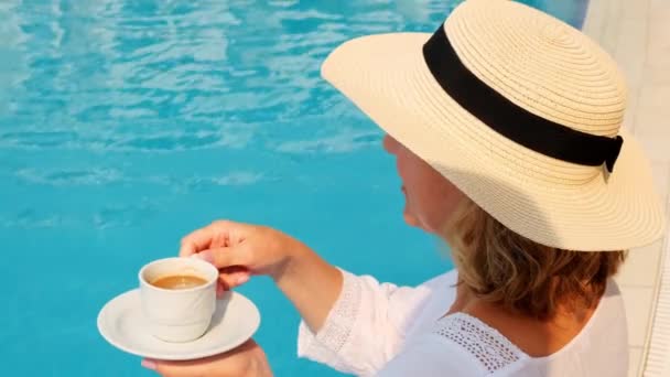 Primo piano di una donna di 50-55 anni con un cappello di paglia che beve da una tazza di caffè accanto a una piscina blu, lussuoso buongiorno, inizio giornata — Video Stock