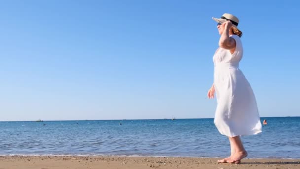 Mujer madura con sombrero de paja y vestido blanco disfrutando del sol caminando a lo largo de la costa azul del mar en un día soleado de verano, disfrutando de la libertad y la relajación. El concepto de un adulto mayor la vida cotidiana — Vídeos de Stock