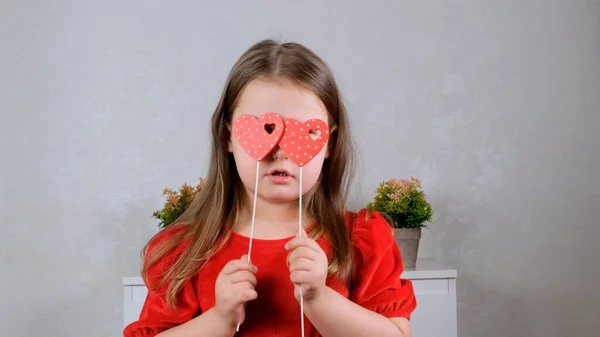 Drôle Jolie petite fille dans une robe rouge avec deux cœurs sur un bâton ferme les yeux avec eux. Concept de Saint-Valentin. — Photo