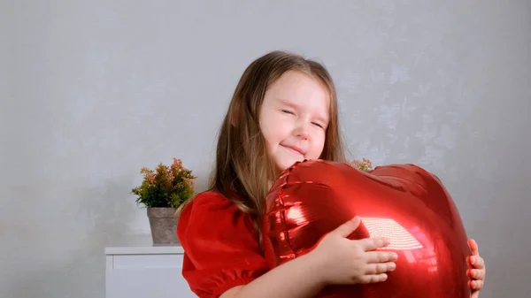 Little cute girl w czerwonej sukience delikatnie przytula czerwone serce w kształcie balonów z jej rąk. Koncepcja Walentynki — Zdjęcie stockowe