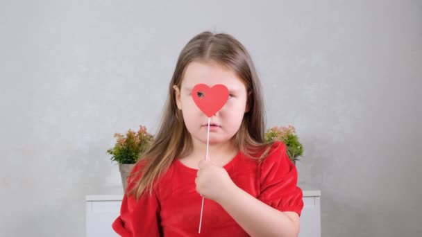 Zabawna dziewczynka w czerwonej sukience z sercem na patyku zamyka oczy. Koncepcja Walentynek. — Wideo stockowe
