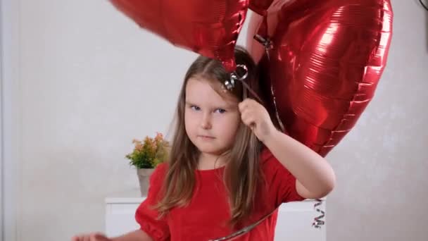 Gadis kecil yang sedih dengan gaun merah dengan tiga balon berbentuk hati untuk hari valentine. Cinta keluarga. hadiah untuk liburan. kebahagiaan dan ciuman — Stok Video