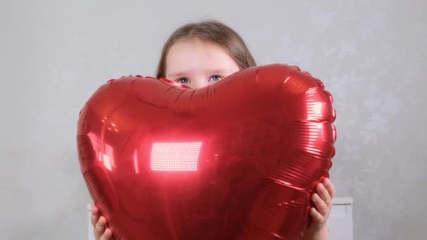Маленькая милая девочка в красном платье нежно обнимает шарики в форме сердца руками. День святого Валентина — стоковое видео