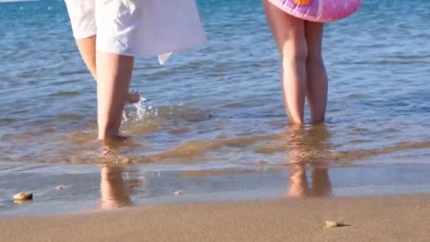 Bedstemor og barnebarn går langs kysten og plasker i havet med fødderne på en varm sommersolskinsdag. Begrebet sommerferie, sjov, familie – Stock-video